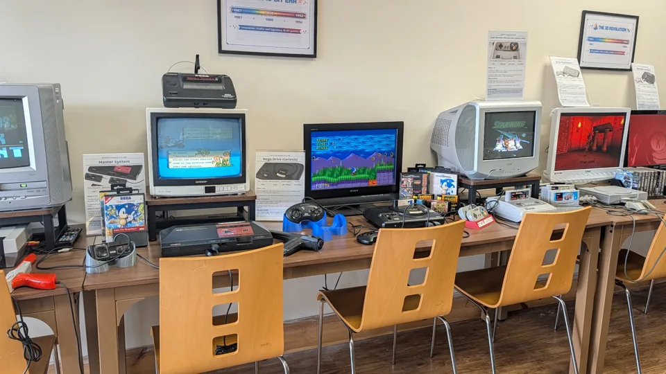 Playable consoles and CRT screens arranged along a row of wooden chairs